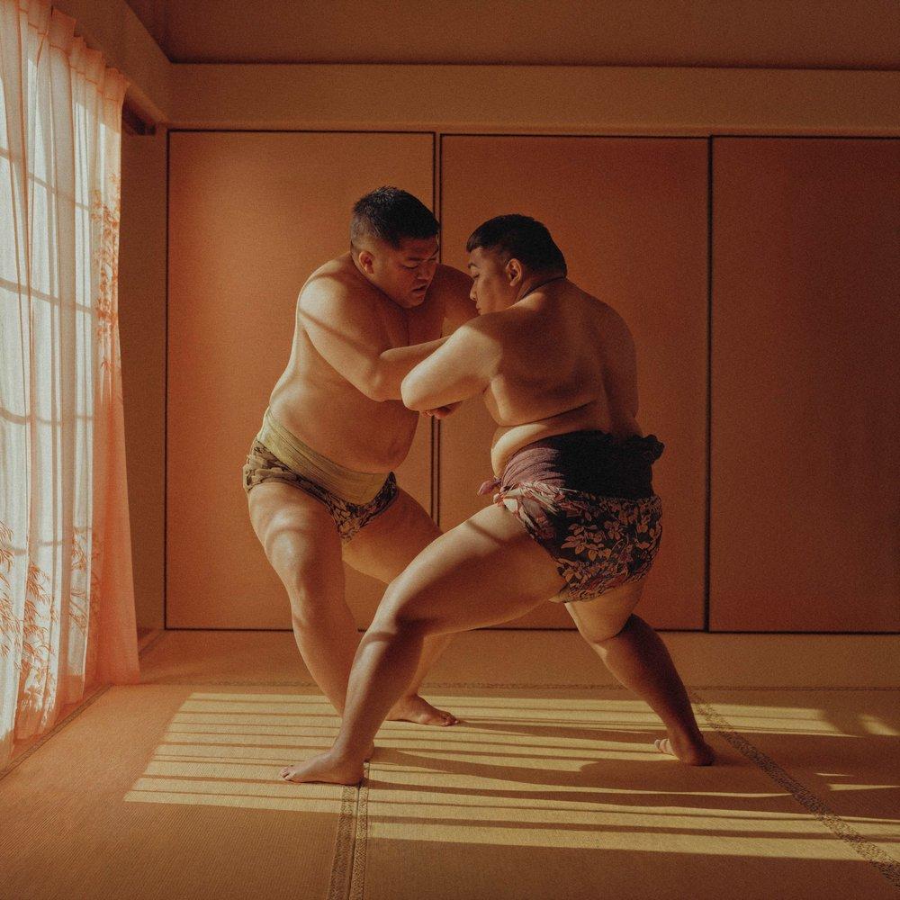 Two men in traditional Japanese loincloths, known as 'mawashi', are engaged in a sumo wrestling match in a dimly lit, traditional Japanese room.
