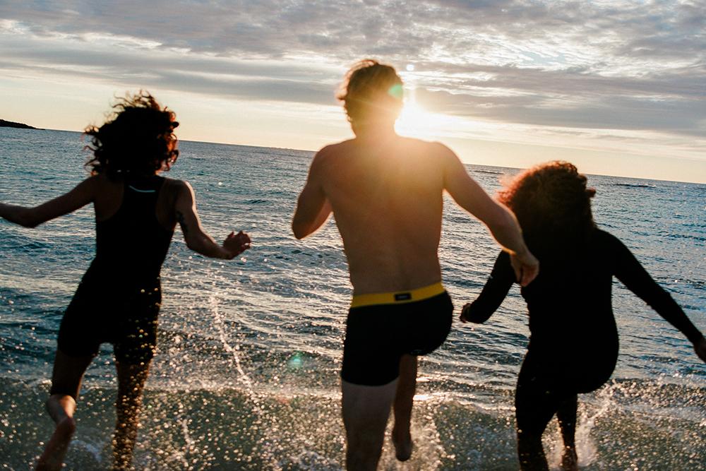 Three people running into the ocean during sunset.