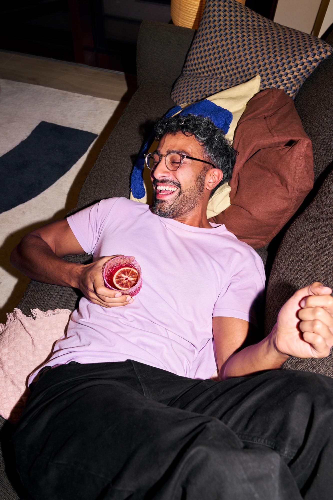 A man in a pink t-shirt and black trousers is sitting on a couch, holding a glass of red liquid with a slice of orange in it.