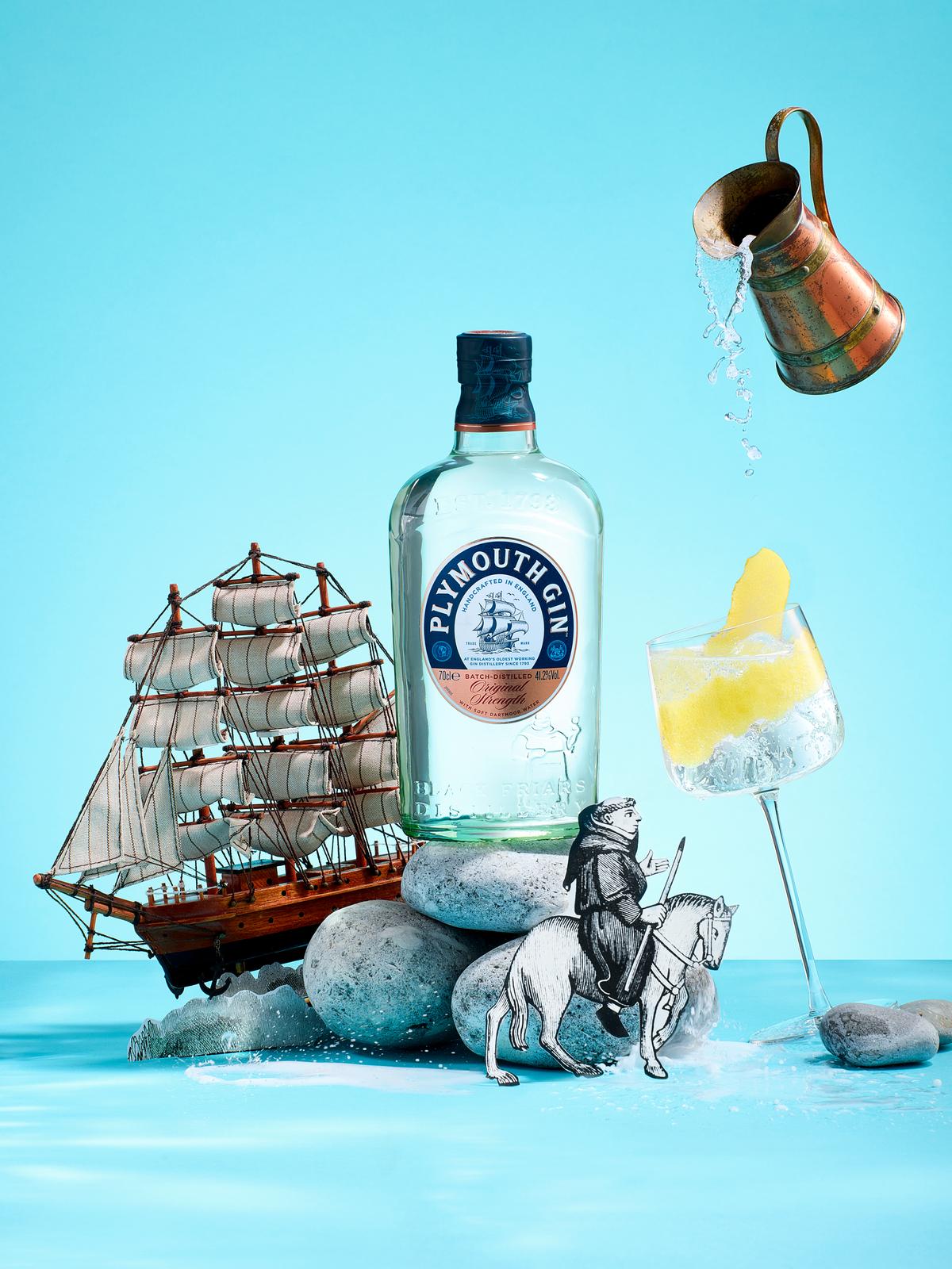 A still life featuring a bottle of Plymouth Gin, a copper jug, a glass of gin and tonic, a ship model, and rocks on a blue background.