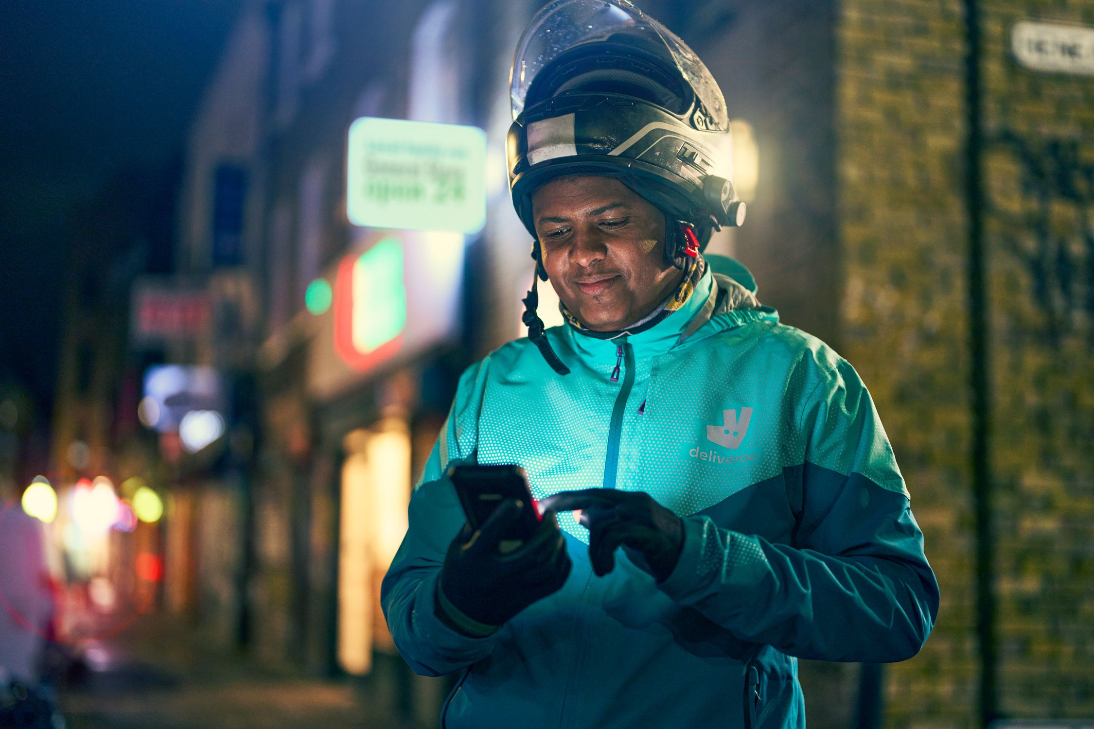 A person in a Deliveroo uniform, wearing a helmet and holding a smartphone, standing in front of a city street at night.