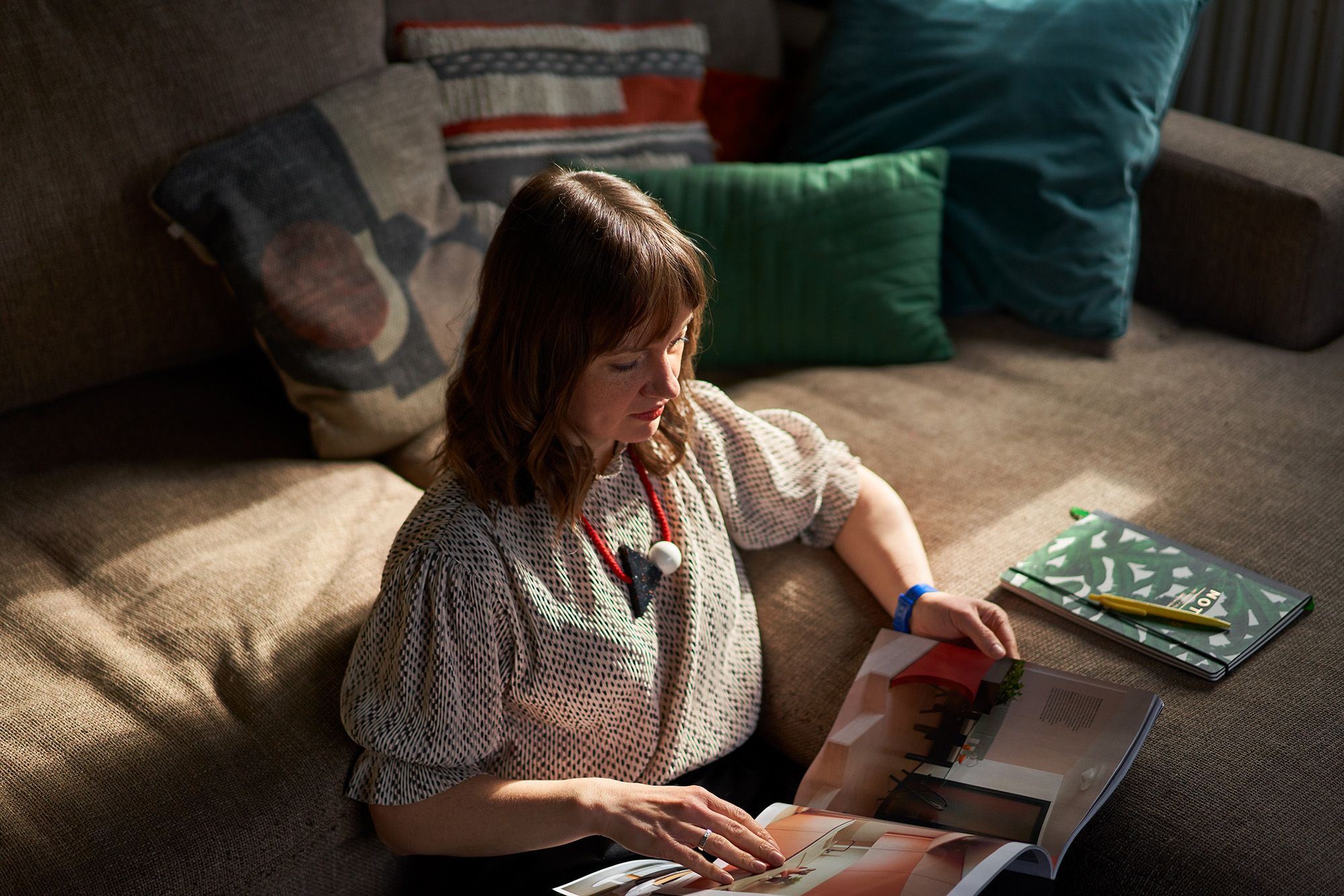 A woman sits on a couch, surrounded by books and papers, looking at a portfolio of her artwork.