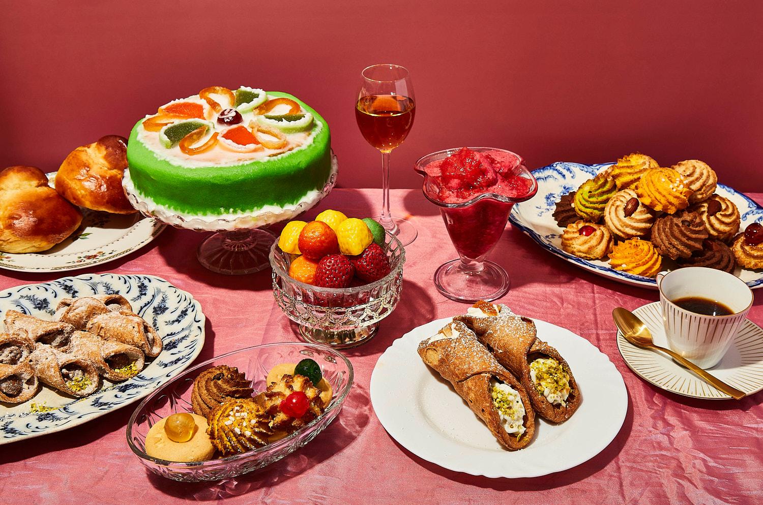 A table set with a variety of Italian desserts, including a green-topped cake, cannoli, and fruit, on a pink tablecloth.