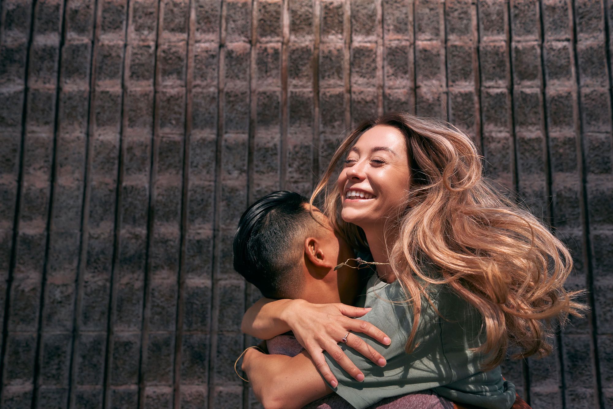 A woman with long, curly brown hair and a green t-shirt is being hugged by a man with short, dark hair and a grey t-shirt, in front of a brick wall.