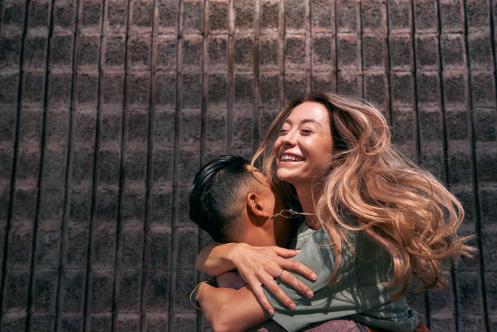 A woman with long, curly brown hair and a green t-shirt is being hugged by a man with short, dark hair and a grey t-shirt, in front of a brick wall.
