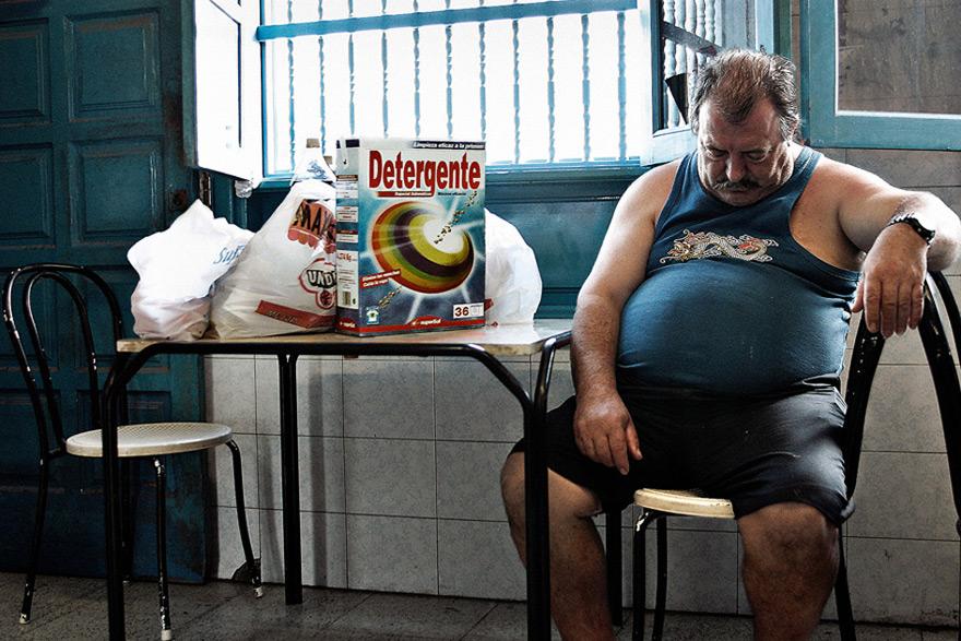 A man in a blue tank top and black shorts is sitting in a chair, surrounded by a table with a few items, including a box of laundry detergent, a bag of food, and a few other items.
