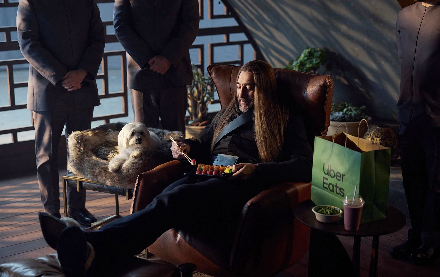 A man with long hair and a black suit sits in a chair, surrounded by a dog, a table with food and a green Uber Eats bag, in a modern, Asian-inspired room with a large window.