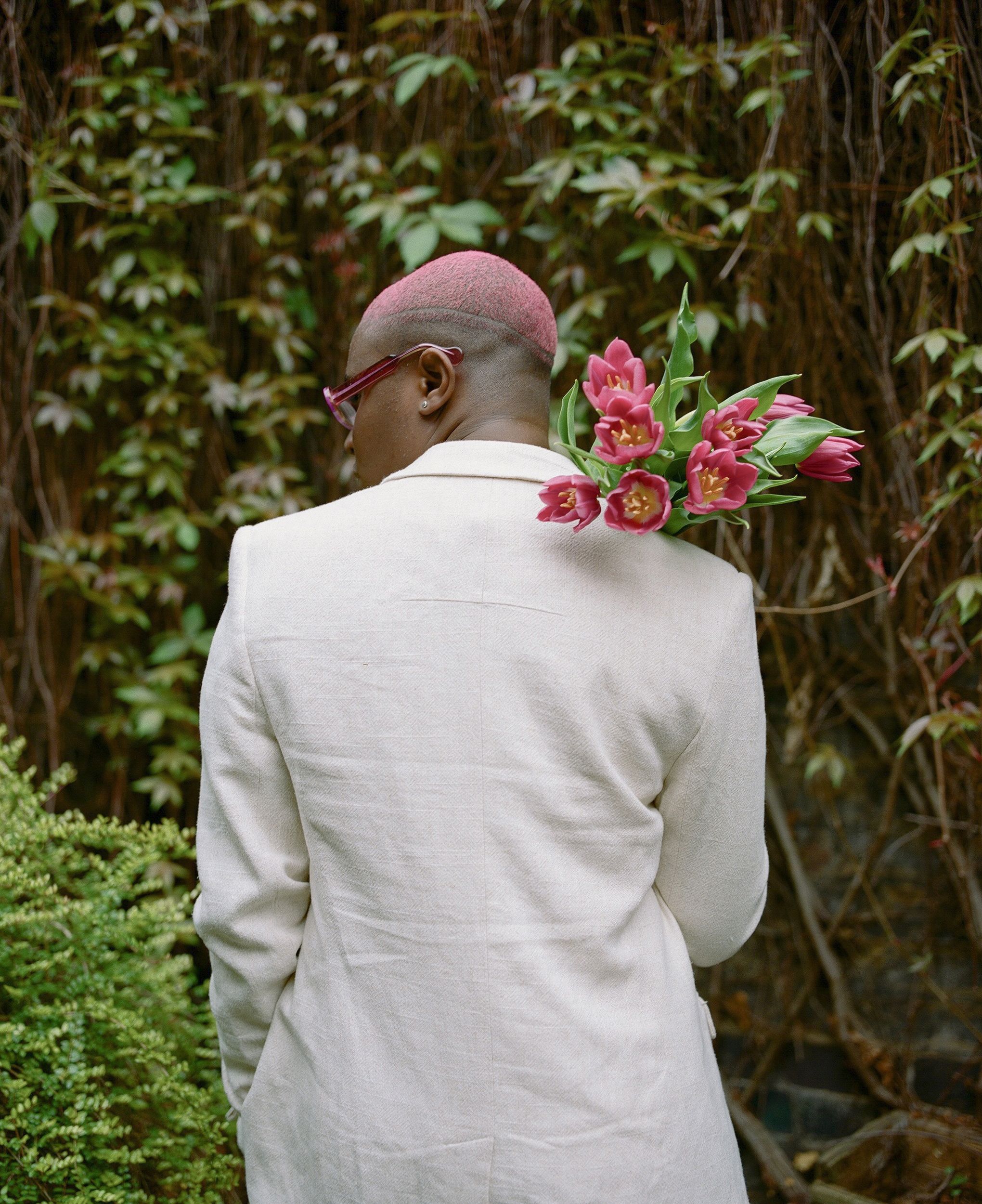 A person with a shaved head and pink hair, wearing a white suit and holding a bouquet of pink tulips, standing in front of a green wall with vines.