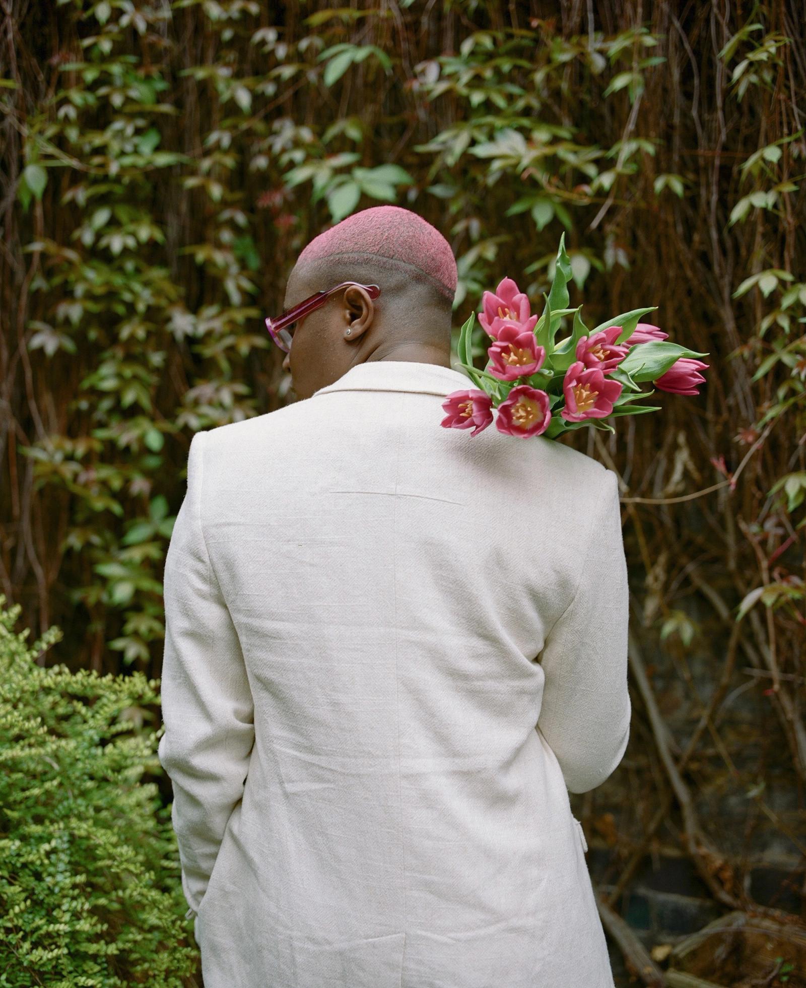 A person with a shaved head and pink hair, wearing a white suit and holding a bouquet of pink tulips, standing in front of a green wall with vines.