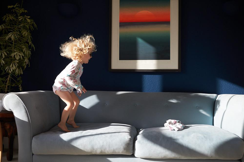 A young girl with curly blonde hair is jumping on a light gray couch in a living room with dark blue walls. She is wearing a long-sleeved white shirt with a colorful pattern and.
