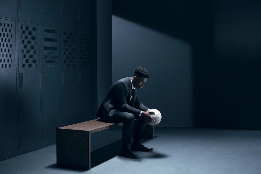 A man in a suit and tie sitting on a bench in a dimly lit locker room, holding a soccer ball.