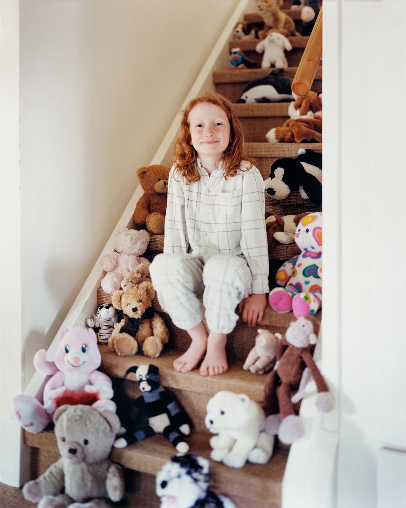 A young girl with red hair sits on a staircase surrounded by a large collection of stuffed animals and toys.