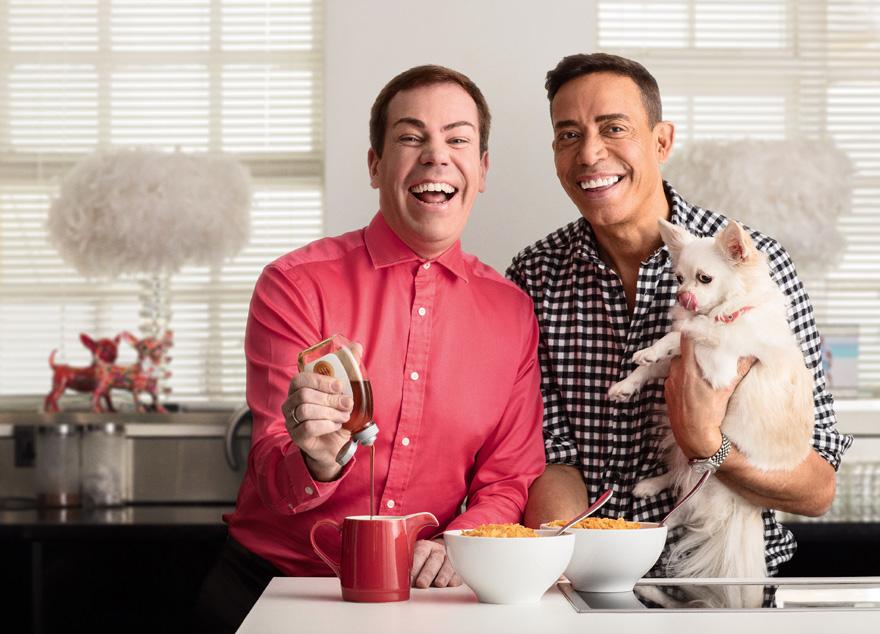 Two men, one holding a small white dog, sitting at a table with a breakfast spread, including cereal, milk, and a red mug.