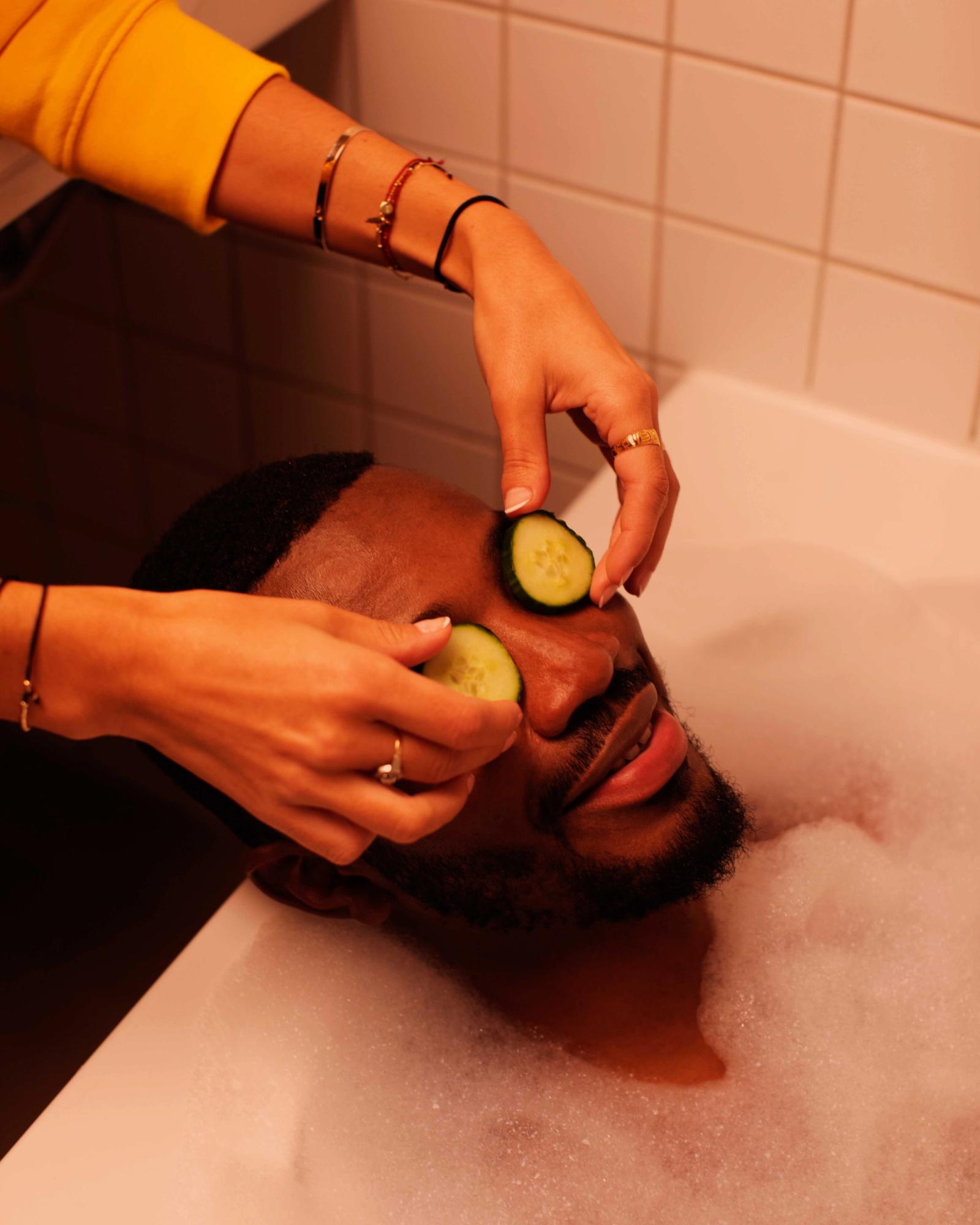 A person, likely a man, is being given a facial treatment in a bathtub, with someone else applying cucumber slices to their eyes.