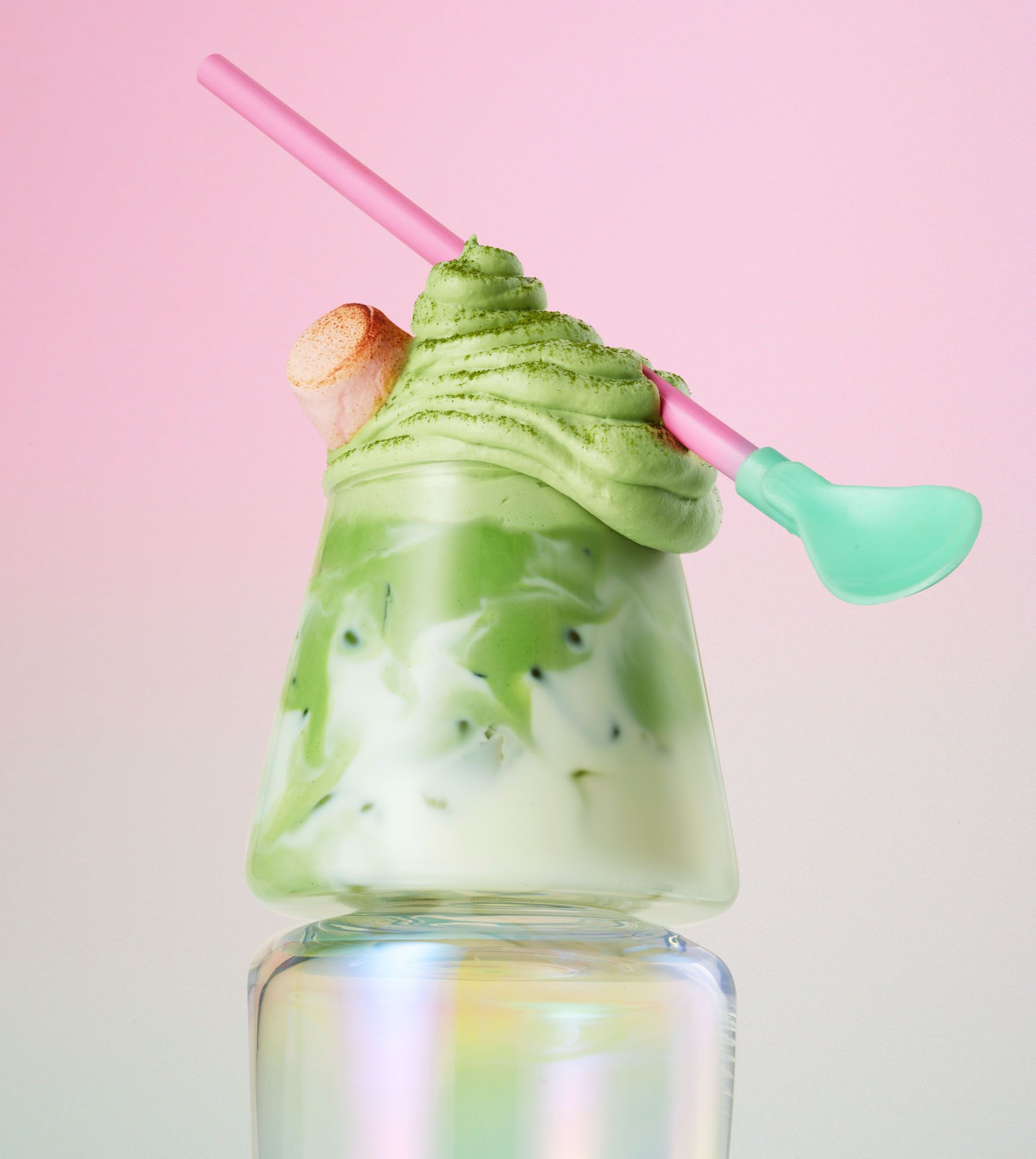 A glass of green, creamy, sweet dessert drink, topped with whipped cream and a pink straw, served in a tall, clear glass.