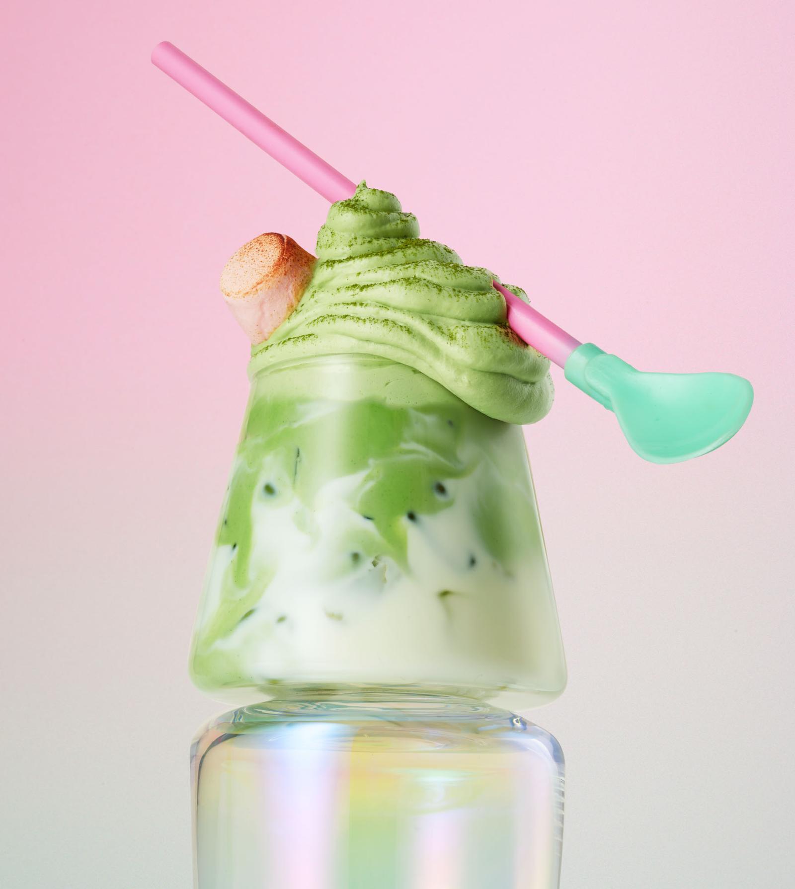 A glass of green, creamy, sweet dessert drink, topped with whipped cream and a pink straw, served in a tall, clear glass.