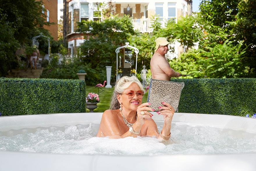 A woman in a hot tub in a garden, holding a leopard-print clutch, with a man in the background mowing the lawn.