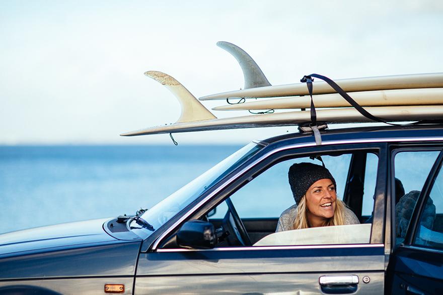 A woman in a black beanie and a grey sweater is sitting in the driver's seat of a car, with surfboards on the roof.