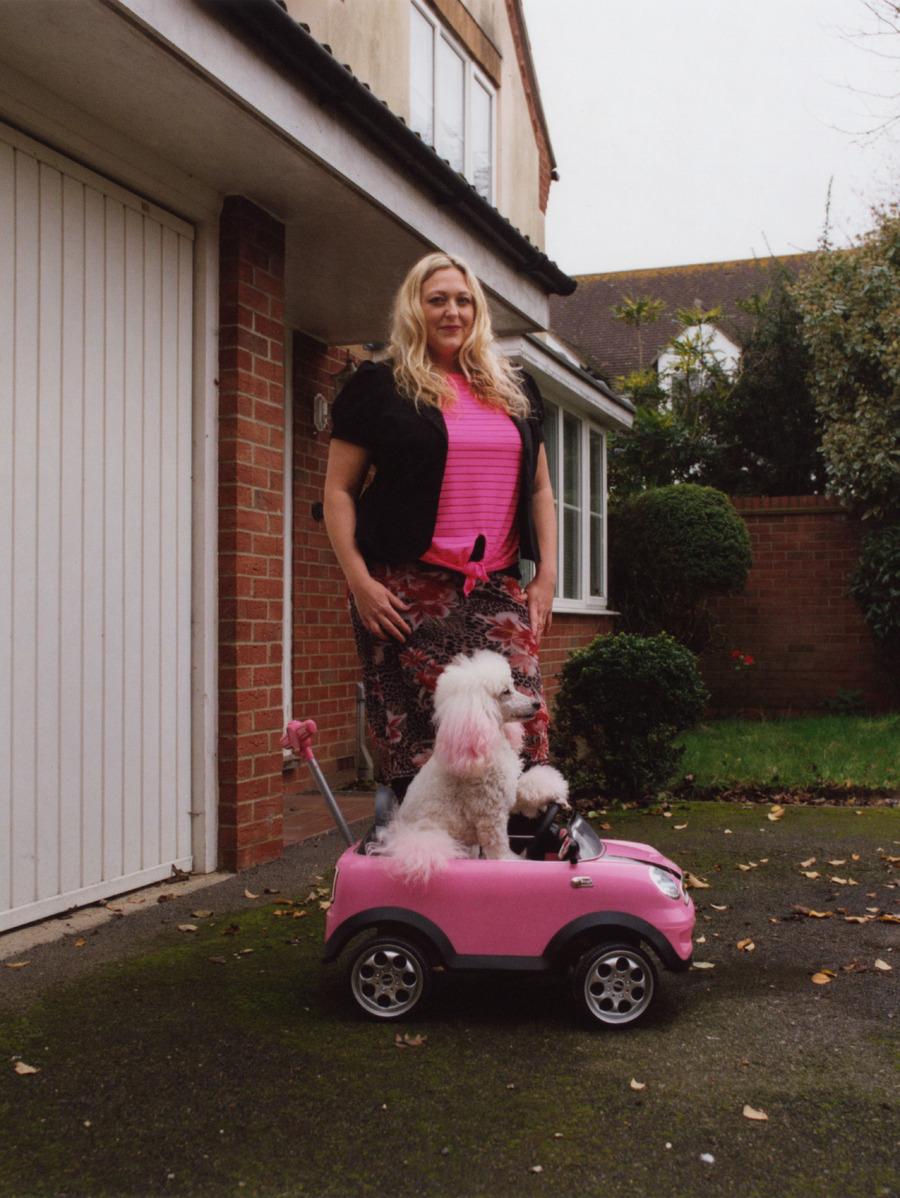 A woman stands in front of a red brick house with a white poodle beside her.
