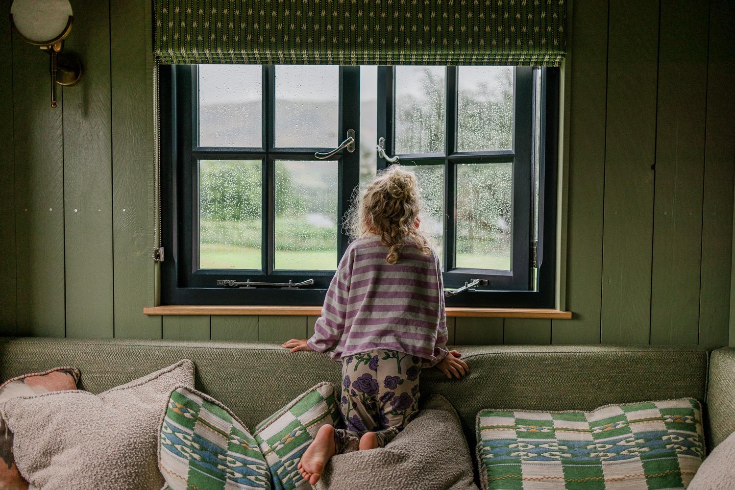 A young girl with curly blonde hair and a striped shirt is standing on a couch, looking out the window at a rainy day.