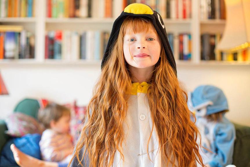 A young girl with long red hair wearing a penguin onesie in a living room with other children in the background.
