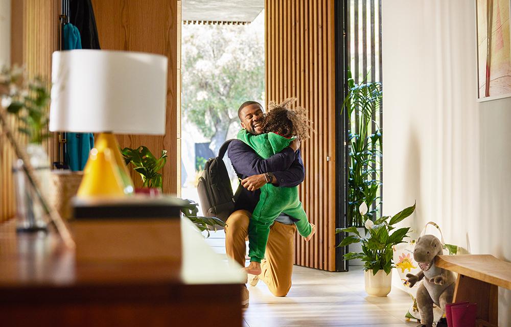 A man in a dark blue shirt and brown trousers is holding a young girl in a green dinosaur onesie in a modern living room.