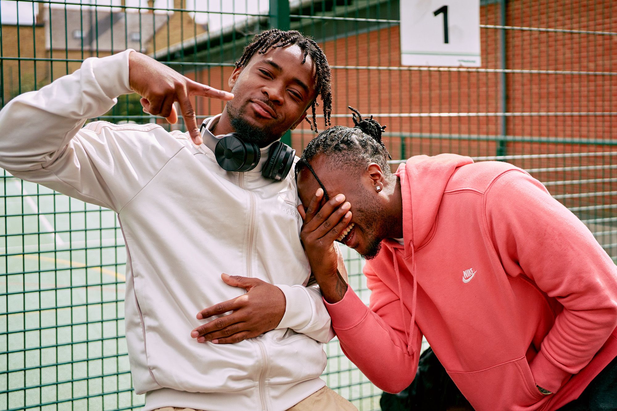 Two men pose casually, with one covering his face, in front of a green fence.