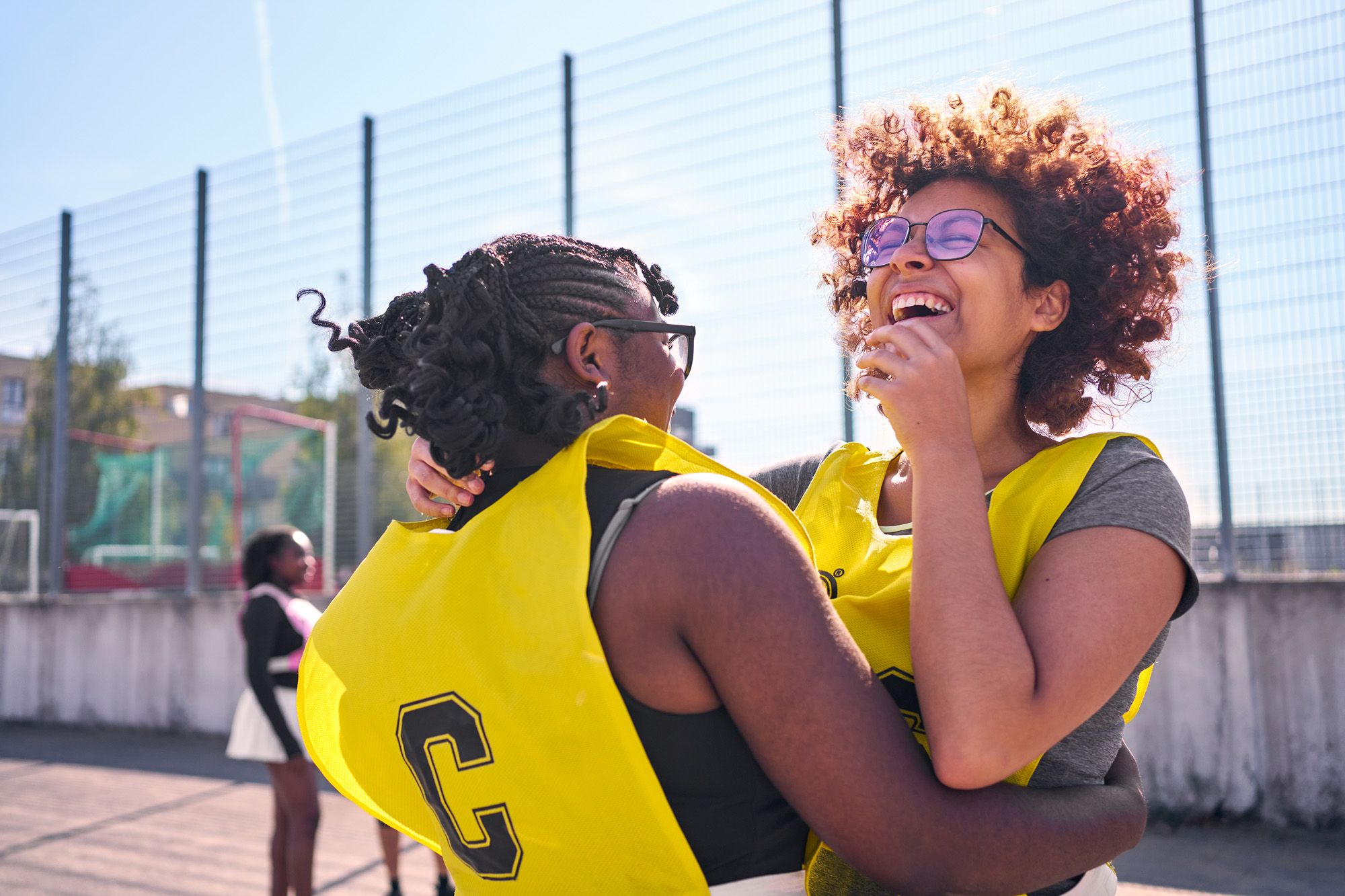 Two women, both with curly hair, wearing yellow bibs with a black 'C' on the back, hugging each other on a basketball court.