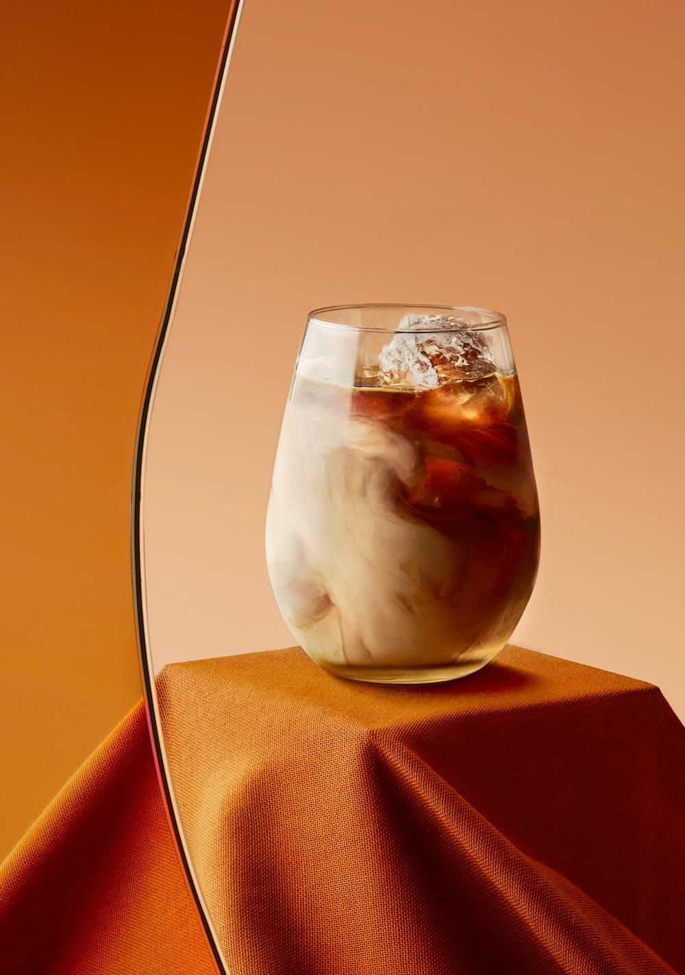 A glass of iced coffee with a brown liquid and ice cubes, served in a clear glass on a tablecloth.