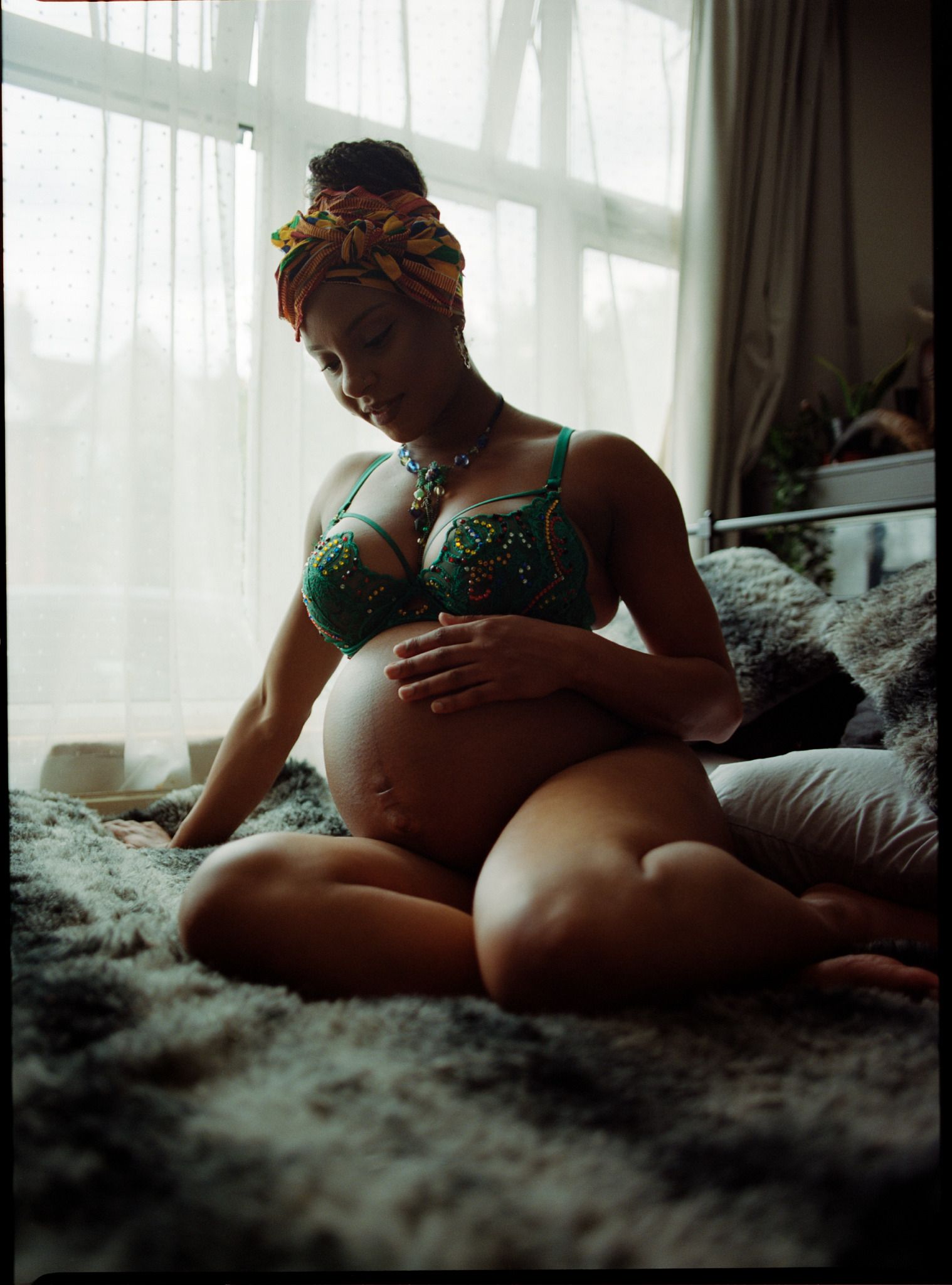 A woman in her early 30s sits on a bed, cradling her pregnant belly with one hand and stroking her own belly with the other. She's wearing a green bra and is lying on a bed with.
