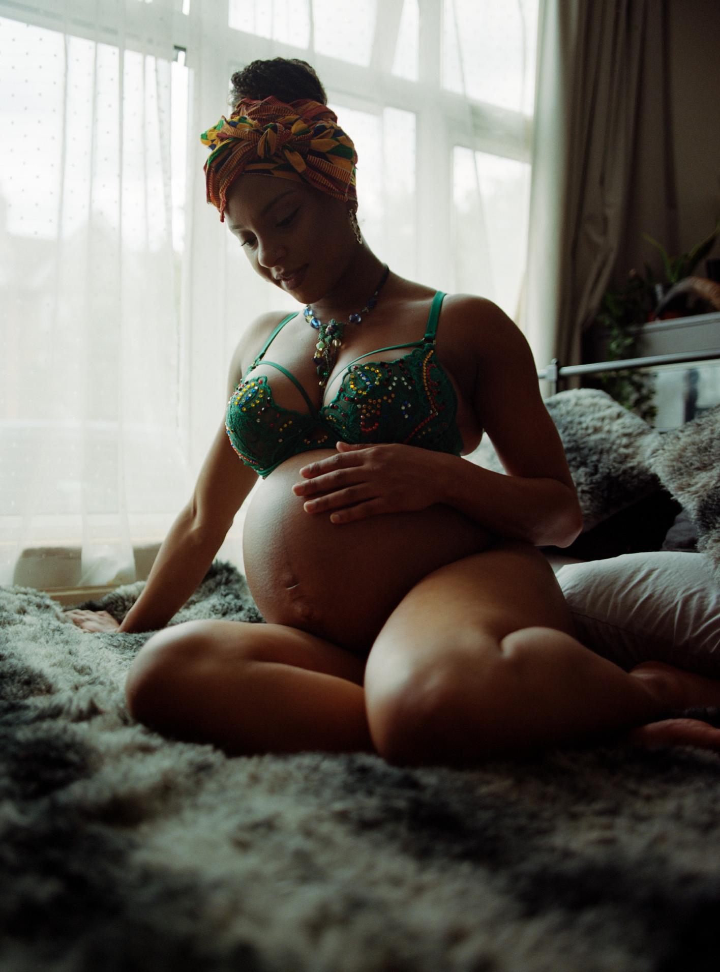 A woman in her early 30s sits on a bed, cradling her pregnant belly with one hand and stroking her own belly with the other. She's wearing a green bra and is lying on a bed with.