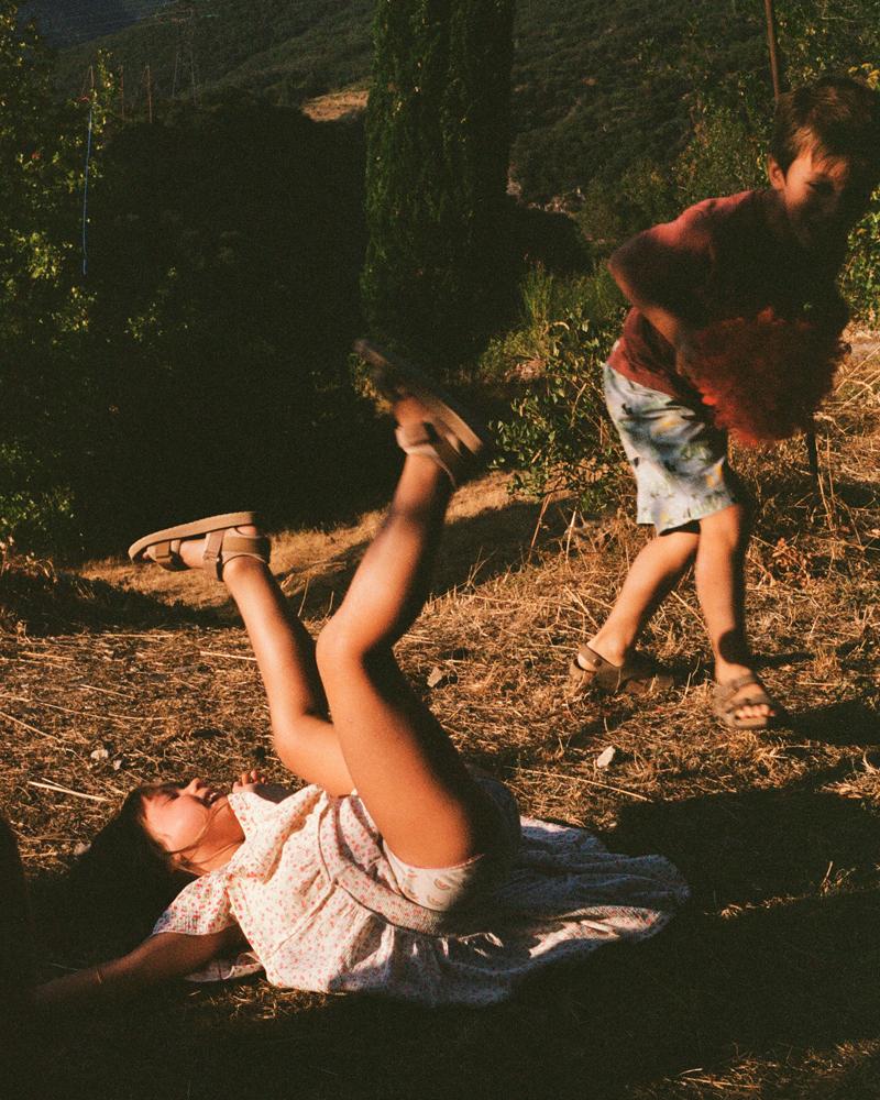 A woman in a white dress with red flowers is lying on the ground, with a man in a red shirt and blue shorts standing over her, in a field with trees and hills in the background.