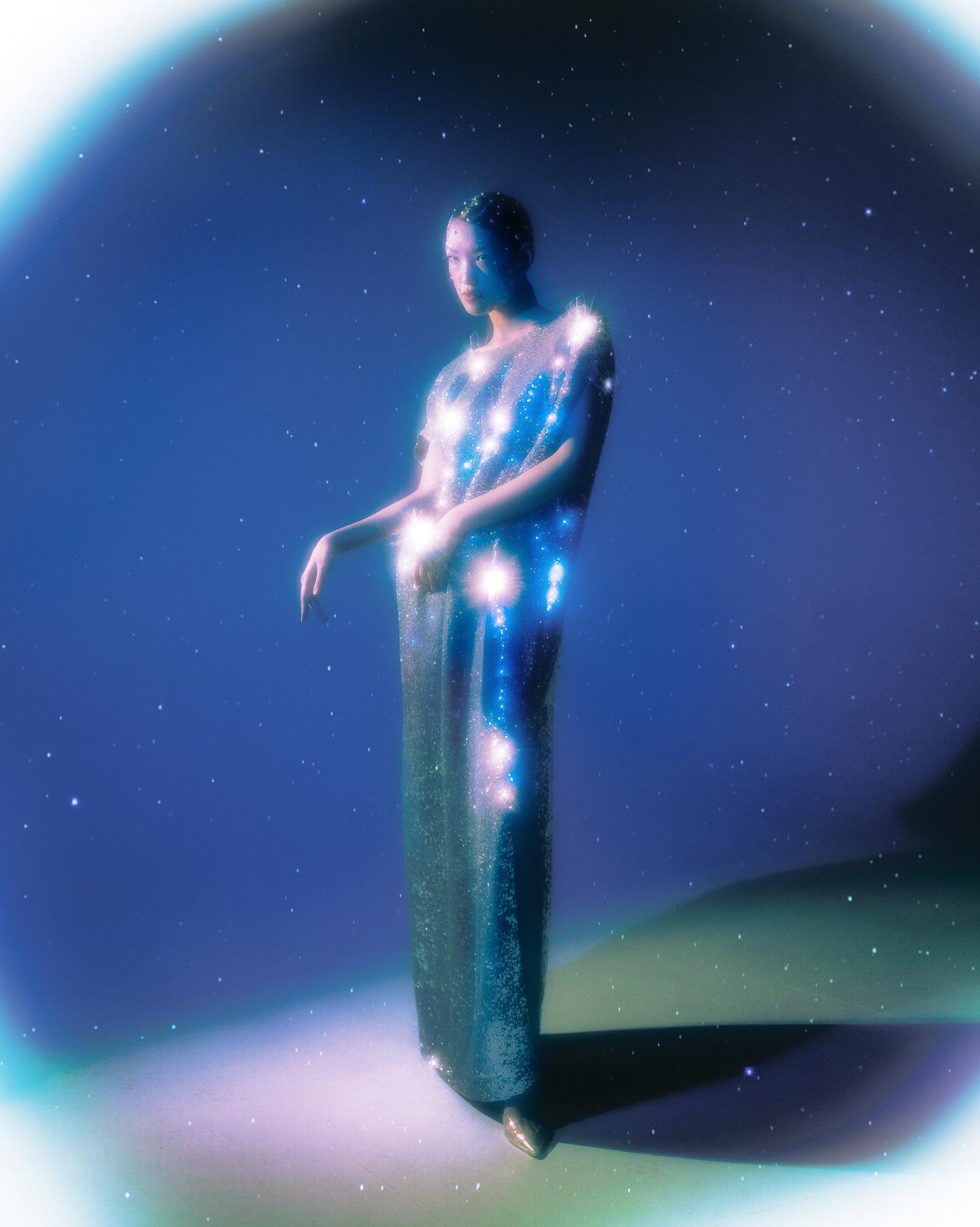 A woman in a long, flowing, star-studded gown, surrounded by a halo of light, standing in a dark, starry night sky.