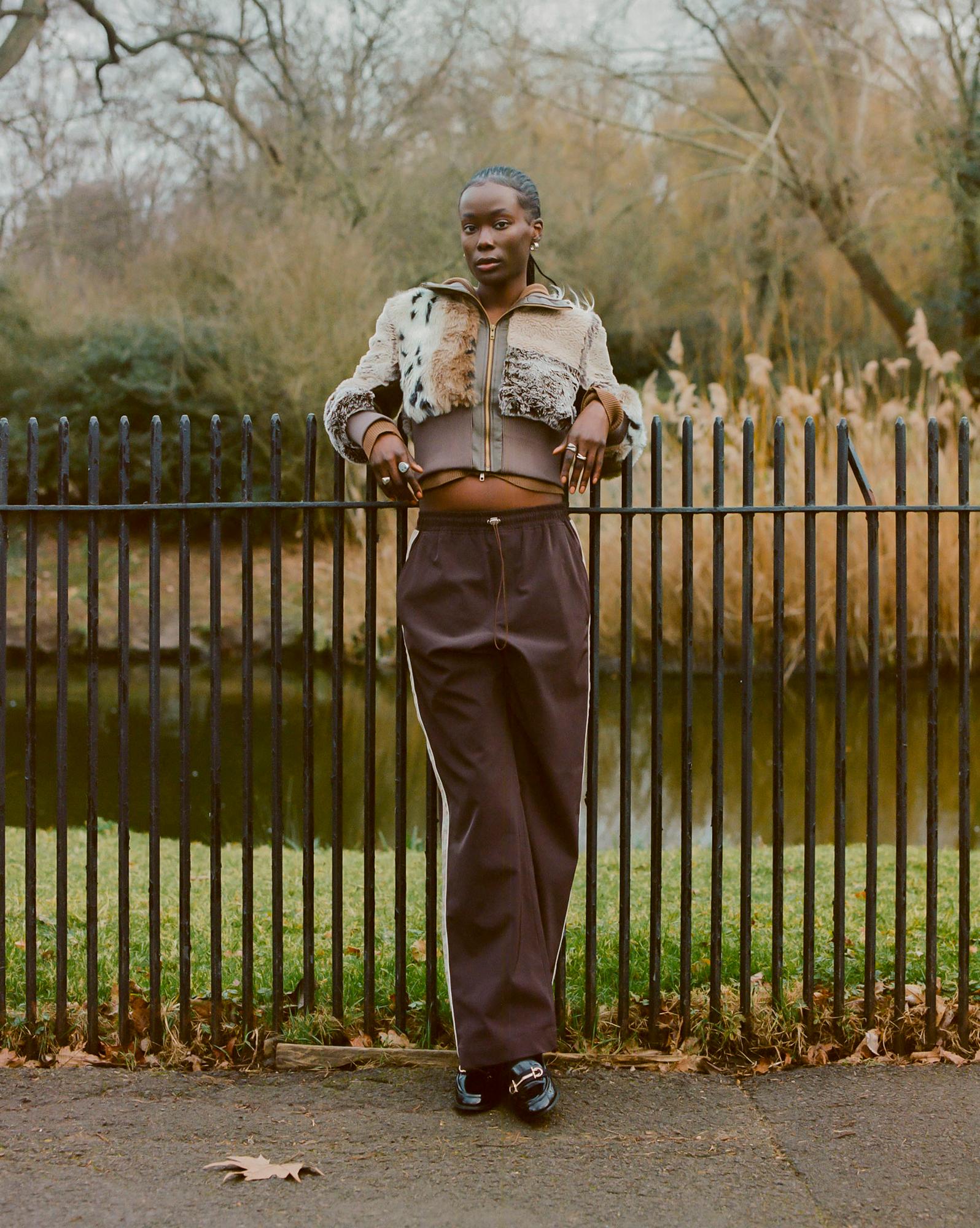 A person with dark skin and braided hair, wearing a fur jacket, a brown sports bra, and brown track pants, standing in front of a black metal fence in a park-like setting.