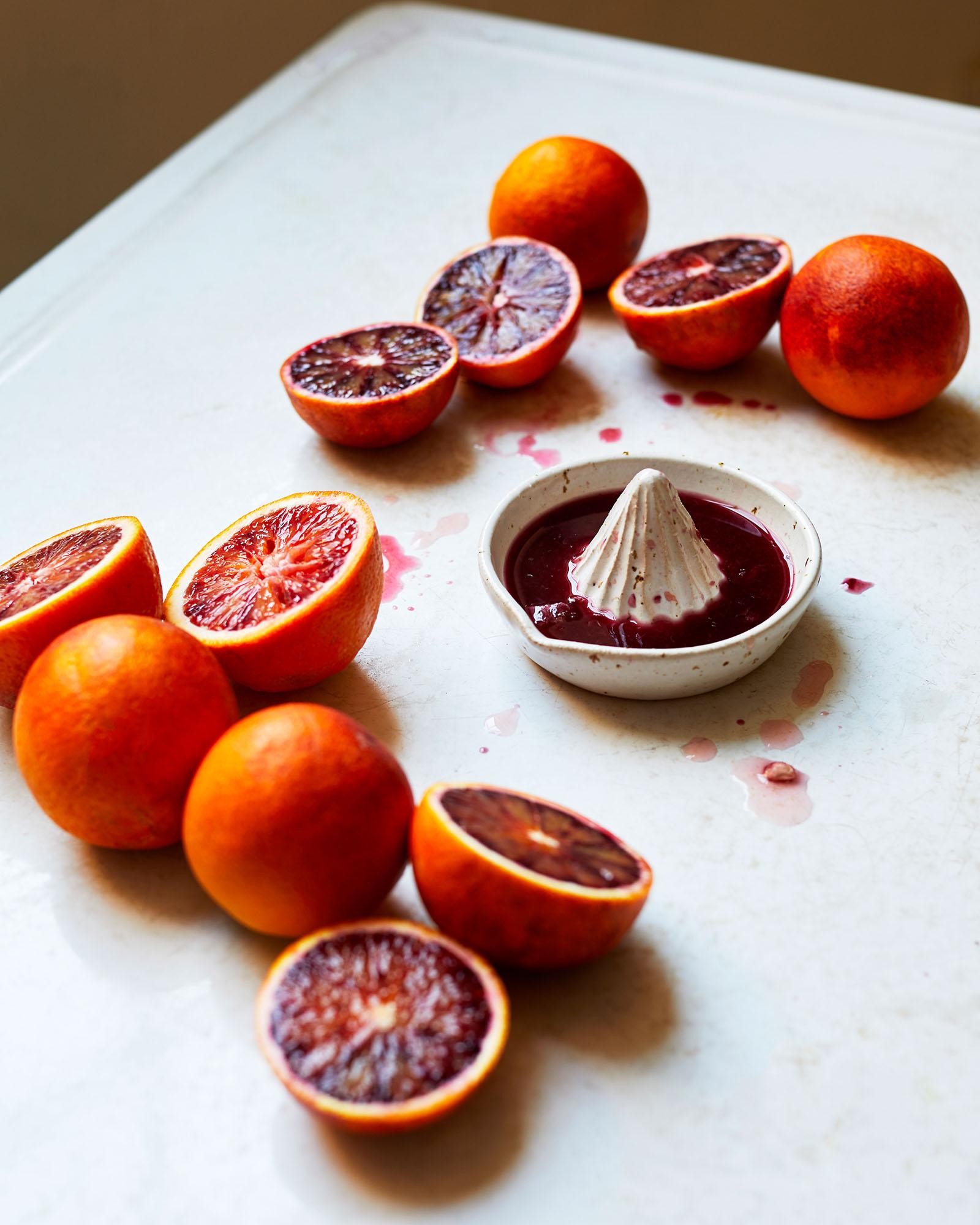 A still life of blood oranges and a ceramic citrus juicer, with some oranges halved and others whole, on a white surface.