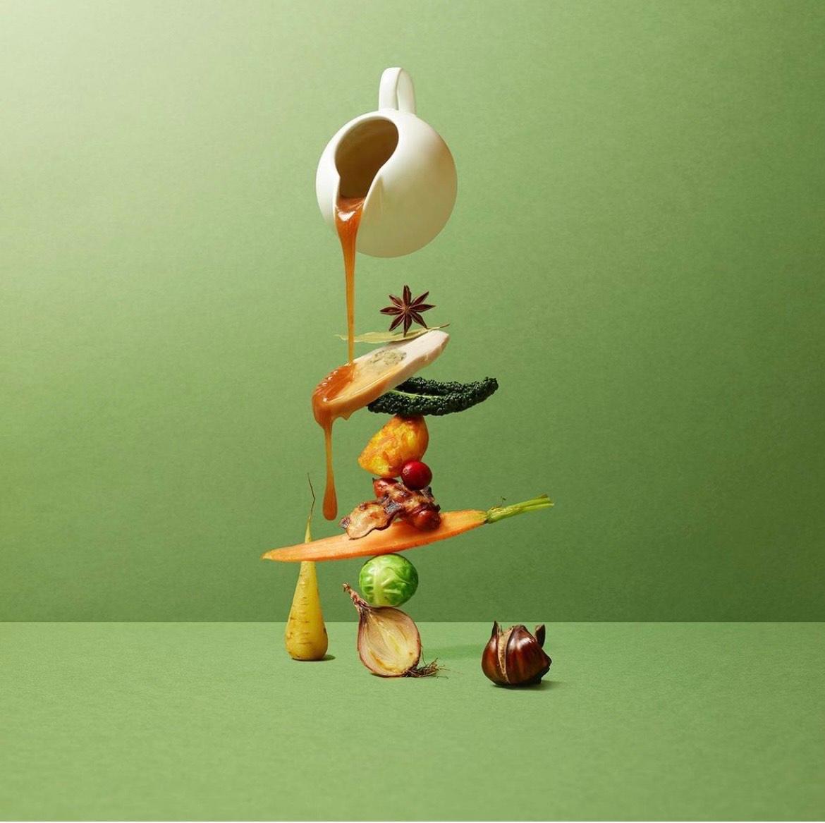 A stack of various food items including a carrot, broccoli, cherry, cheese slice, meat piece, bread slice, and star anise with white sauce pouring from above.
