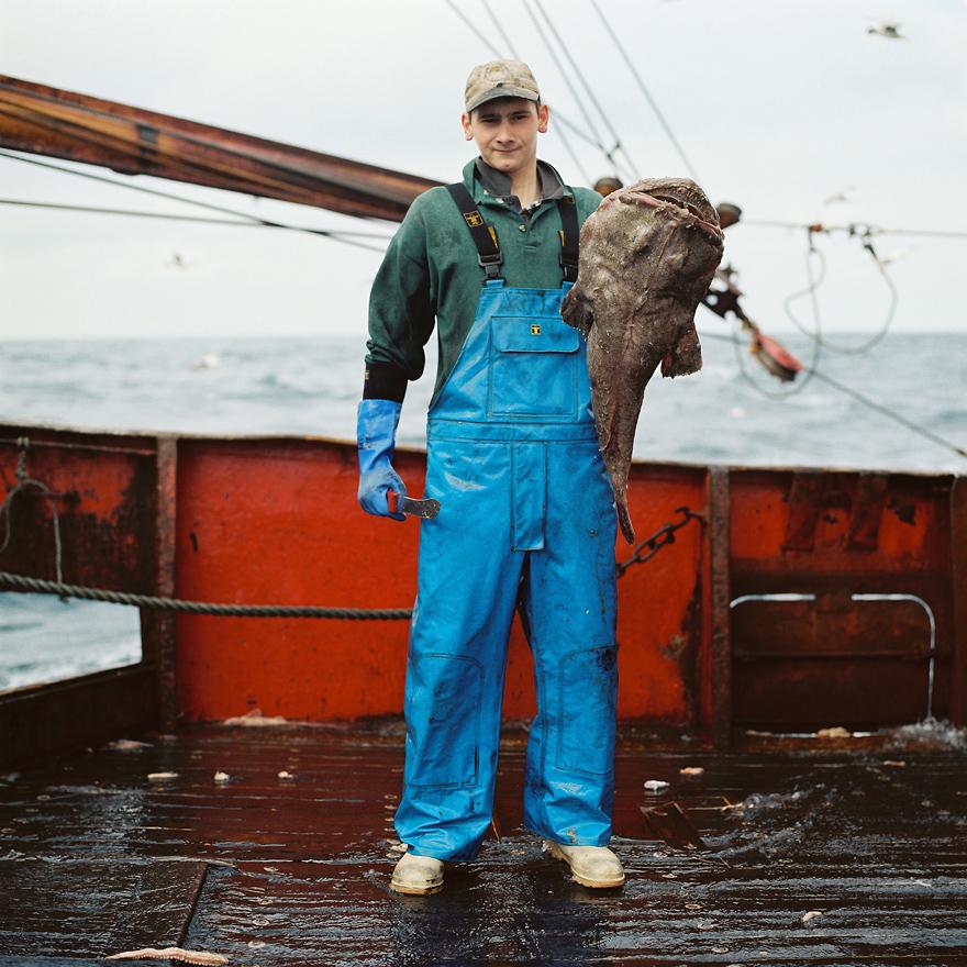 A man in a green sweater, blue overalls, and a baseball cap is standing on a fishing boat, holding a large fish.