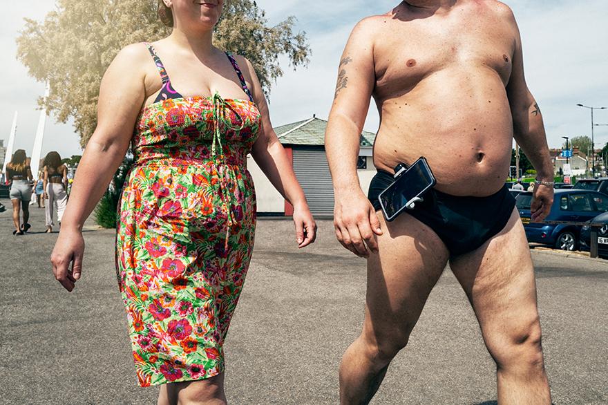 A man in a pair of swimming shorts, walking in a public area, with a woman in a floral dress, walking alongside him.