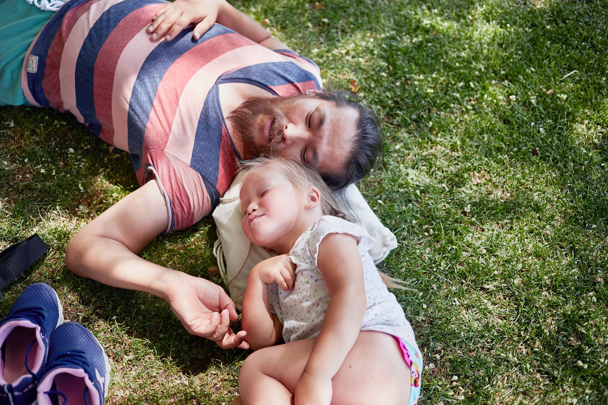 A man and a young girl are sleeping on the grass, surrounded by a few scattered leaves and a small backpack.