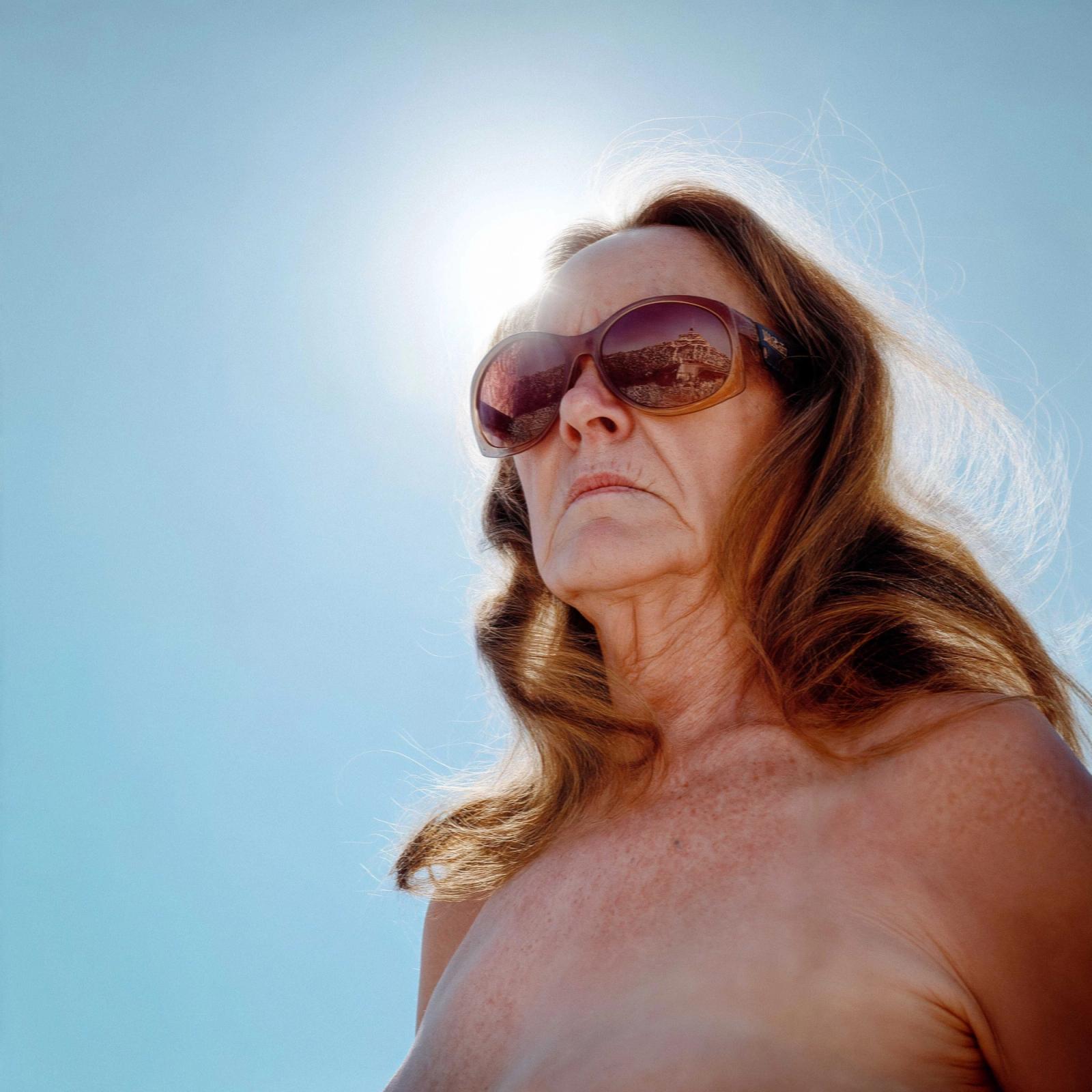 A woman with long, dark hair and a bare, sun-kissed torso, wearing sunglasses and standing in a bright, sunny environment, with a beach or coastal landscape in the background.