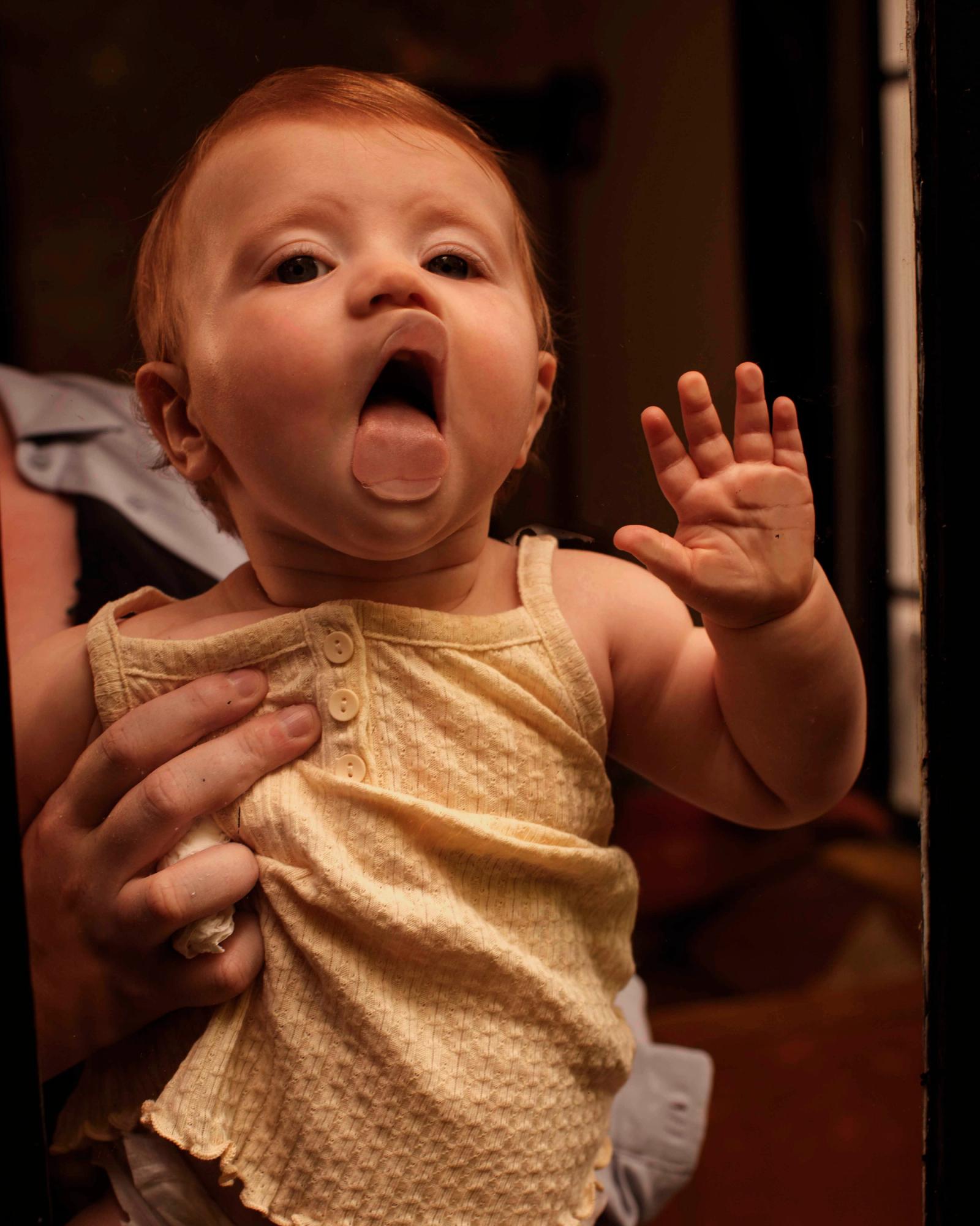 A baby girl with red hair and a yellow tank top is making a silly face and sticking out her tongue.