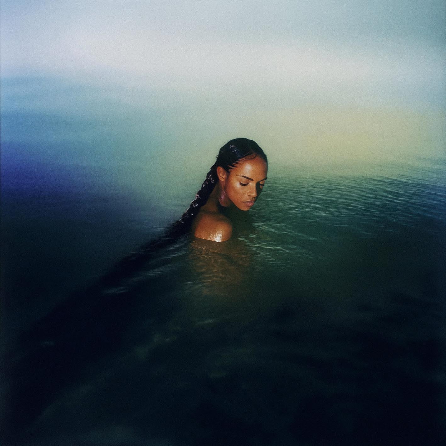 A woman with long, dark hair and a braided ponytail is submerged in water, with her face and upper body visible above the surface.