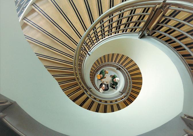A group of people in a spiral staircase, with a metal railing and a large window in the background.