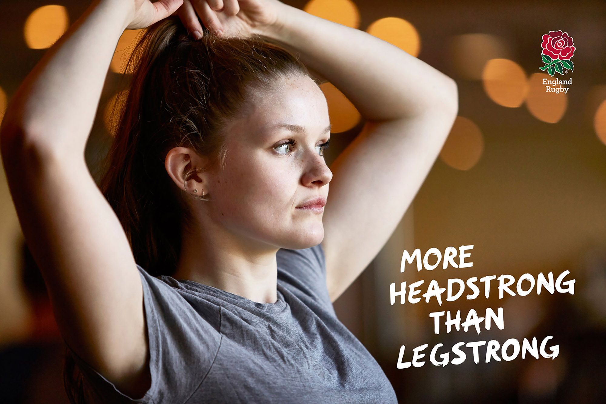 A woman with long hair, wearing a grey t-shirt, is adjusting her hair in a dimly lit room.