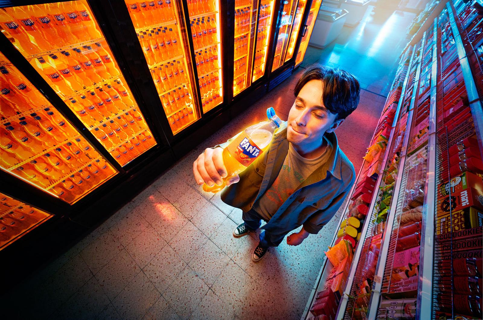 The image shows a man standing in a grocery store aisle, captured from a high-angle "fish-eye" perspective. He is wearing a green jacket and is holding a bottle of orange Fanta up toward his face with a satisfied expression. To his left is a large bank of refrigerated coolers filled with glowing orange bottles of Fanta.