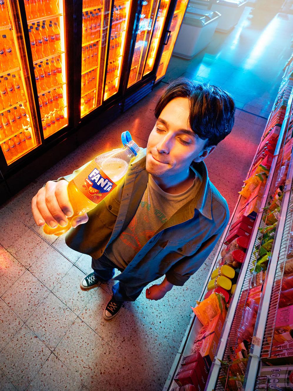 The image shows a man standing in a grocery store aisle, captured from a high-angle "fish-eye" perspective. He is wearing a green jacket and is holding a bottle of orange Fanta up toward his face with a satisfied expression. To his left is a large bank of refrigerated coolers filled with glowing orange bottles of Fanta.