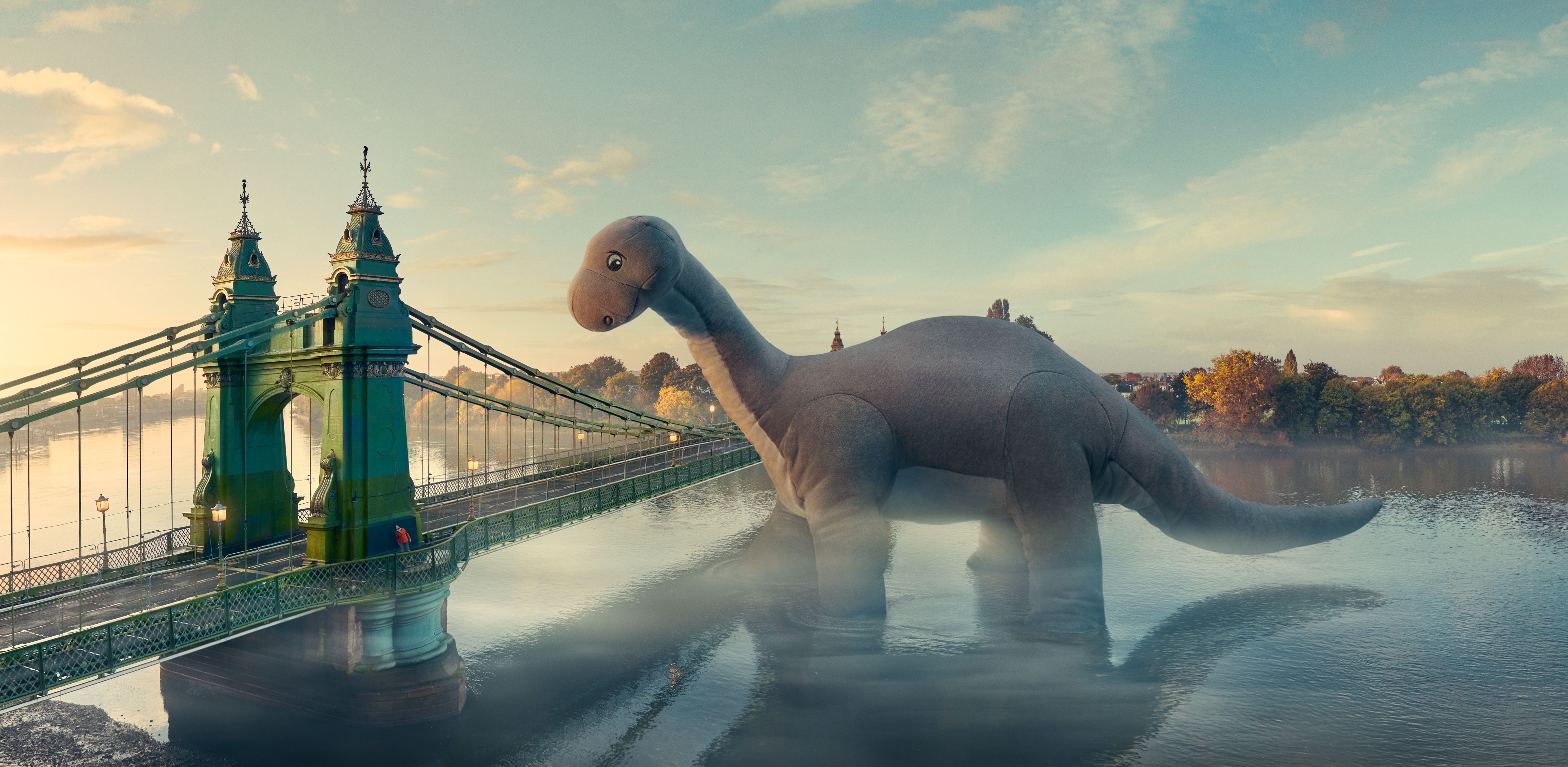 A large, green, metal bridge in london with a central section that is suspended from two towers, with a road and a pedestrian path on either side, and a city skyline in the background and a dinosaur toy enlarged.
