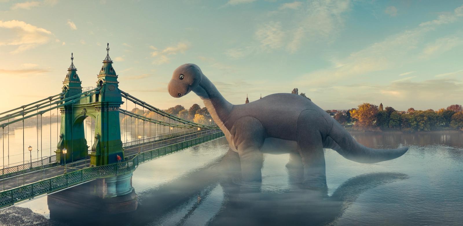 A large, green, metal bridge in london with a central section that is suspended from two towers, with a road and a pedestrian path on either side, and a city skyline in the background and a dinosaur toy enlarged.