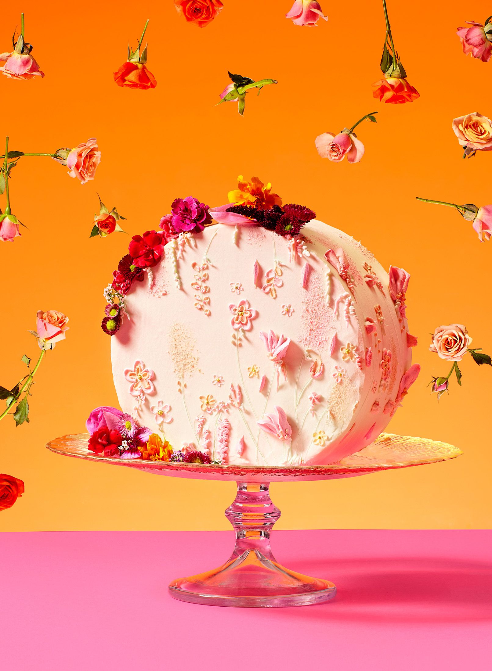 A stunning, multi-layered, pink and white cake with intricate, edible flower decorations, sitting on a glass cake stand on a pink table.