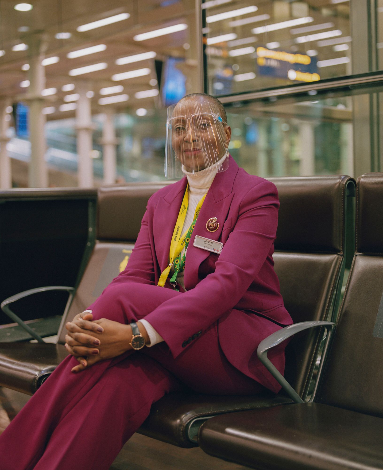 A woman in a magenta suit and face shield sitting on a brown leather chair in an airport or train station.